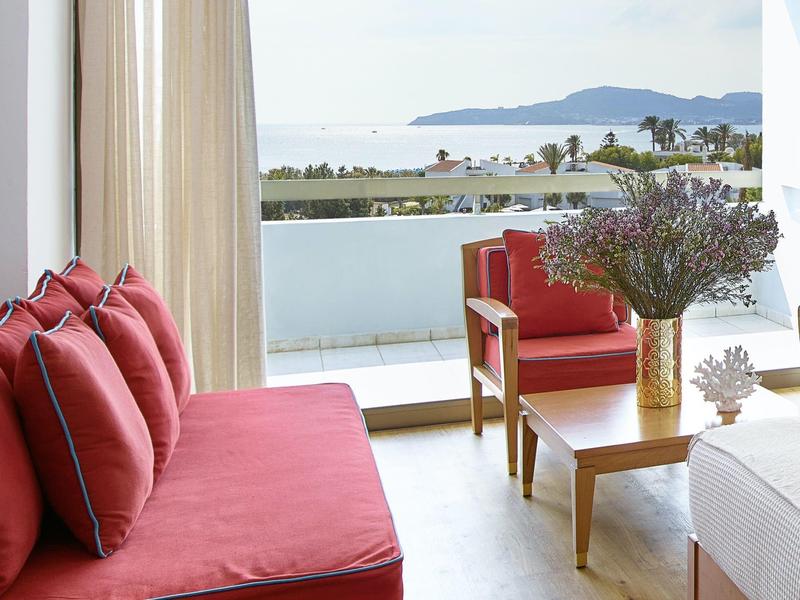 Balcone con vista mare, divano rosso e due poltrone, tavolo in legno con vasi di fiori.