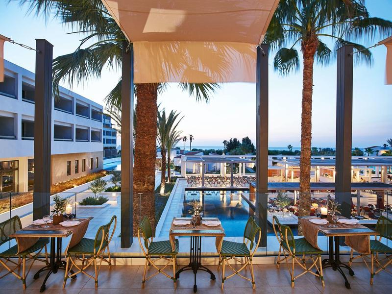 Area esterna moderna di un hotel con tavoli, sedie e piscina sotto palme.