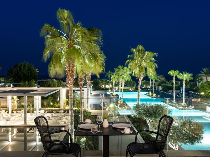 Piscina esterna illuminata con palme e tavolo apparecchiato per due in un ambiente alberghiero di lusso di notte.