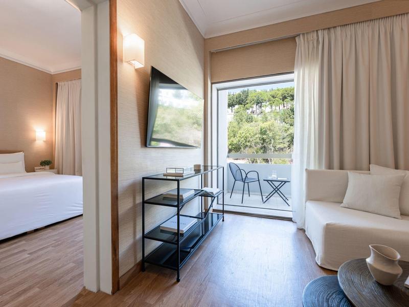 Camera d'hotel con divano bianco, TV e accesso al balcone con vista sul giardino.