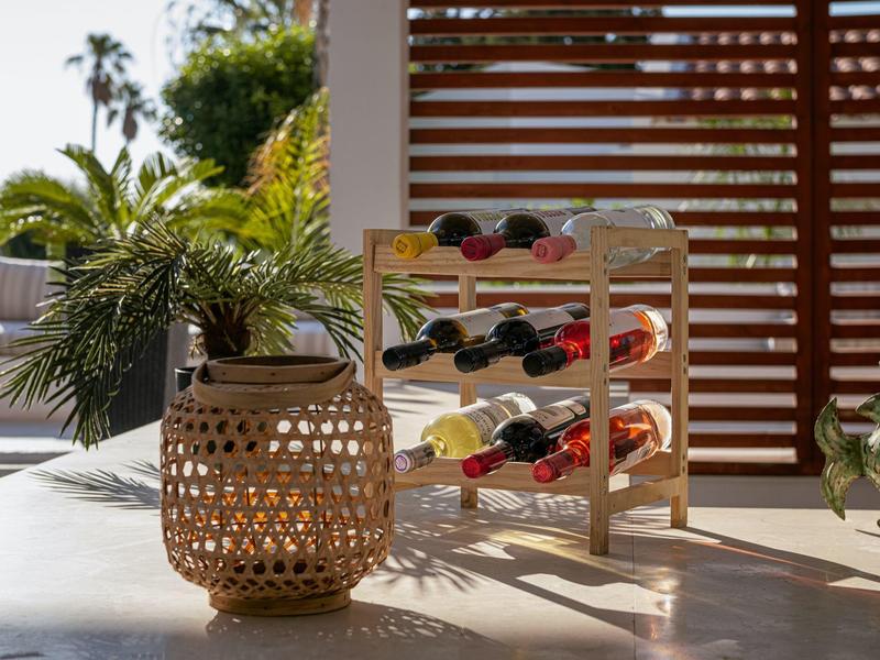 Scaffale in legno per vino con bottiglie accanto a un cesto intrecciato su un tavolo chiaro.