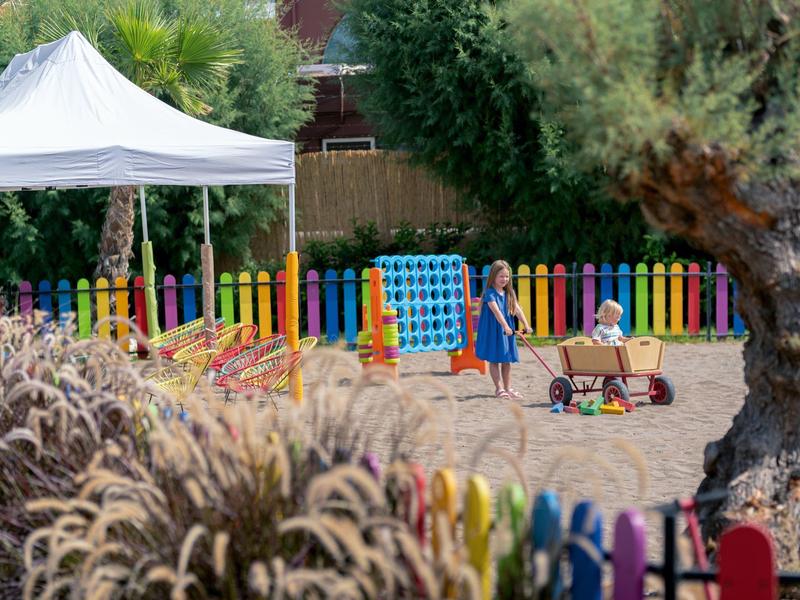 Parco giochi con recinzione colorata, sabbia, strutture per giocare e due bambini sotto una tenda bianca.