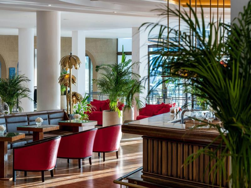 Luminosa hall dell'hotel con poltrone rosse, grandi piante e arredi moderni.
