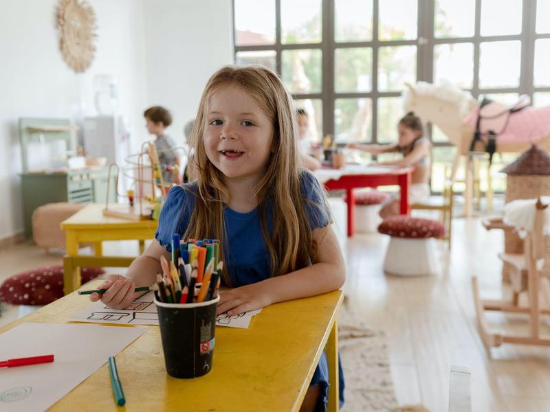 Bambina seduta a un tavolo giallo con matite colorate in una stanza luminosa e accogliente.