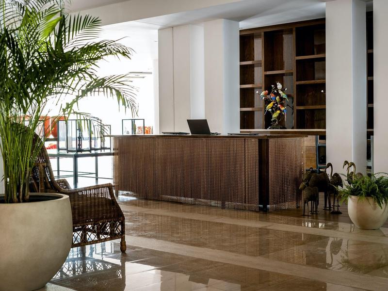 Reception di hotel moderna con banco in legno, area salotto e grandi piante in ambienti luminosi.