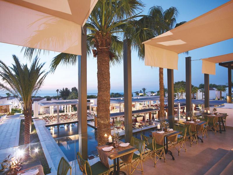Terrazza dell'hotel con tavoli e sedie, palme, piscina e vista sul mare al tramonto.