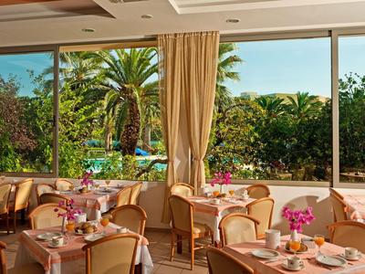 Helles Restaurant mit Fensterblick auf Garten und Swimmingpool, Tische mit weißen Tischdecken und Blumen.