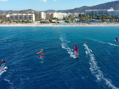 Mehrere Windsurfer fahren vor einem Hotelstrand auf dem tiefblauen Wasser.