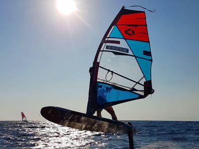 Surfer mit rotem Segel gleitet über das Wasser bei sonnigem Himmel.