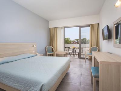 Habitación de hotel luminosa con cama doble, balcón, dos sillas y televisor en la pared.