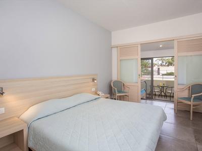 Habitación de hotel luminosa con cama doble, dos sillas y acceso a un balcón con mesa y sillas.