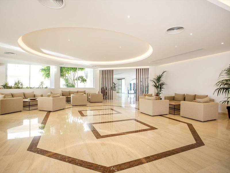 Espaciosa y moderna zona de vestíbulo de hotel con sofás claros, grandes plantas y suelo brillante.