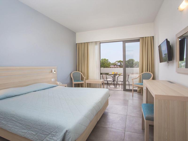 Habitación de hotel luminosa con cama doble, balcón, dos sillas y televisor en la pared.