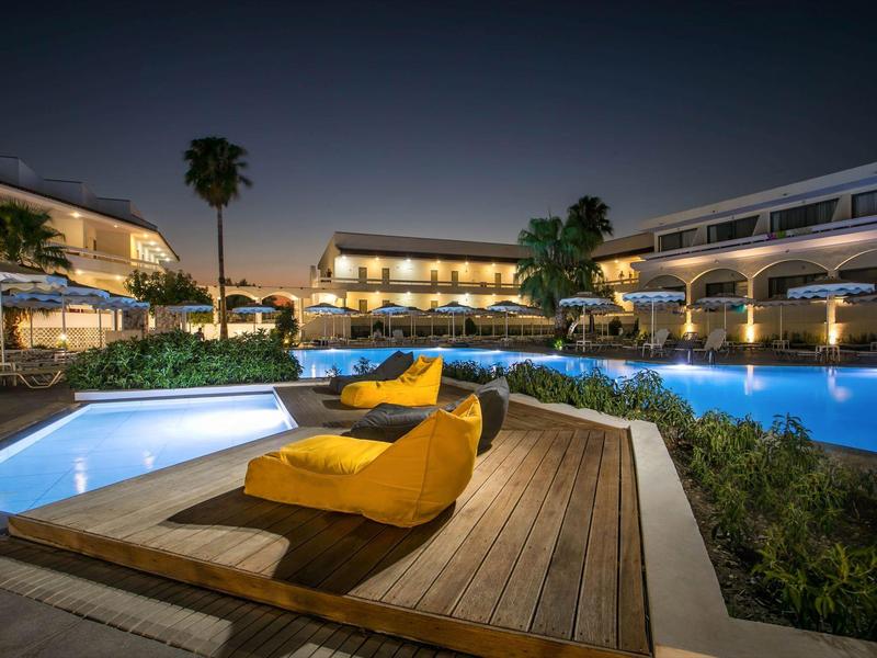 Área de piscina de hotel por la noche con agua iluminada y cojines amarillos en terraza de madera