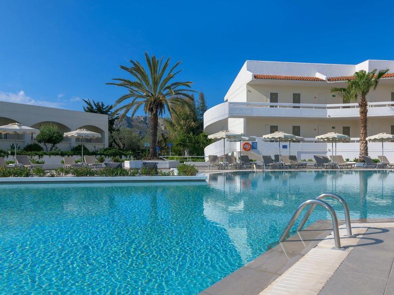Hotel con piscina clara, sombrillas y palmeras bajo cielo azul.