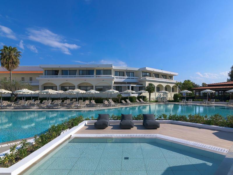 Gran hotel con piscina, tumbonas y sombrillas bajo cielo azul.