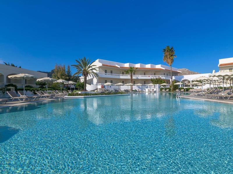Gran piscina de hotel con palmeras y edificios blancos bajo un cielo azul claro.
