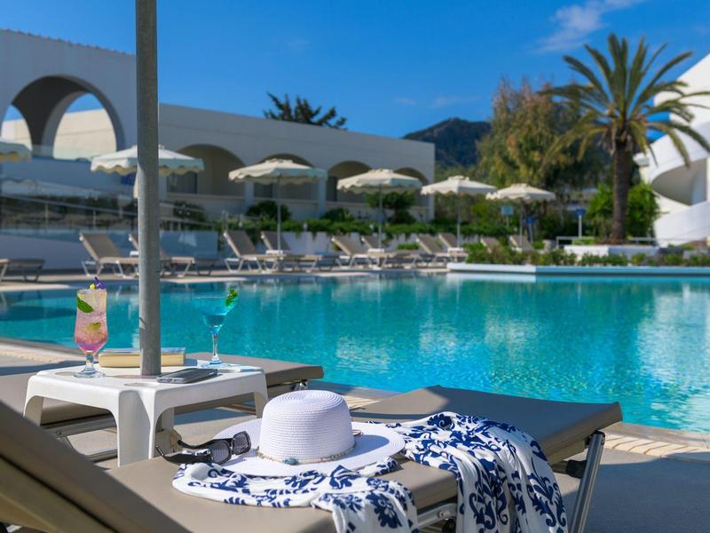 Piscina de hotel con tumbonas, sombrillas blancas y bebida al lado bajo cielo azul claro.