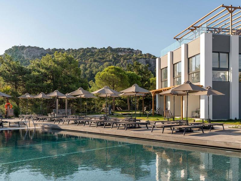 Nowoczesny basen hotelowy z leżakami i parasolami na tle zalesionego wzgórza.
