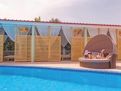 Rundes Lounge-Bett mit Sonnenschirm am blauen Pool vor Haus mit Glasfenstern und Holzverkleidung.
