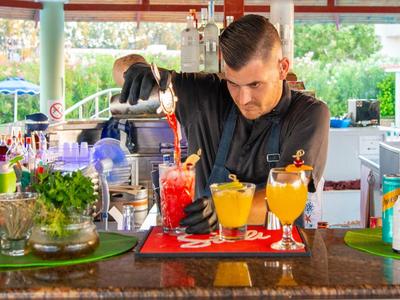 Ein Barkeeper mixt bunte Cocktails an einer sonnigen Bar mit Pflanzen und Getränkeflaschen.
