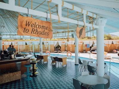 Espace piscine ouvert avec bar, tables et chaises, avec un panneau 'Welcome to Rhodes'.