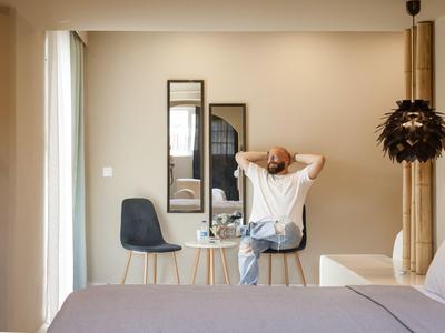 Mann sitzt auf Stuhl am kleinen Holztisch in hellem Zimmer mit großem Bett und dekorativer Lampe.