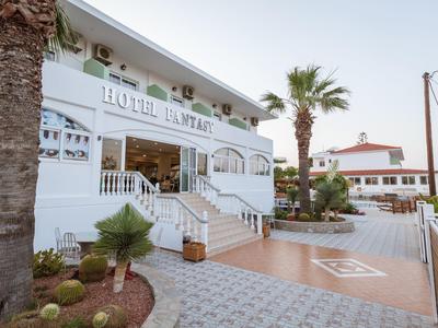 Weißes Hotelgebäude mit Garten, Palmen und Terrasse bei klarem Himmel.