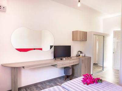 Helles Schlafzimmer mit Holzmöbeln, ovalem Wandspiegel, TV, Sessel und Blumen auf dem Bett.