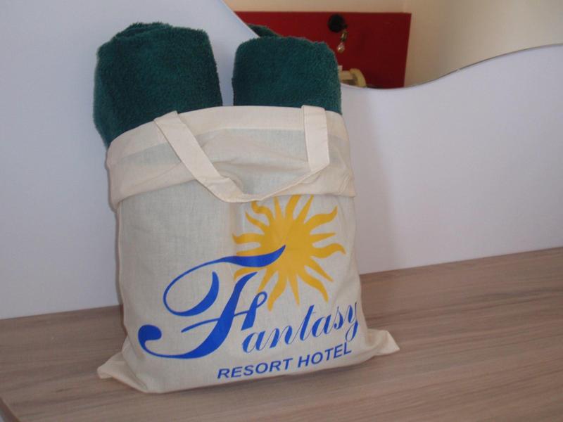 Borsa di stoffa beige con logo Fantasy Resort Hotels e due asciugamani verdi su tavolo di legno.