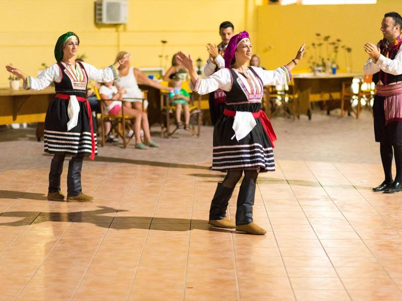 Due donne ballano con abiti tradizionali al chiuso con un uomo che applaude vicino.