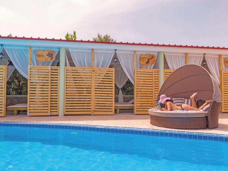 Piscina con acqua blu, accanto a un lettino rotondo davanti a un edificio con persiane in legno.