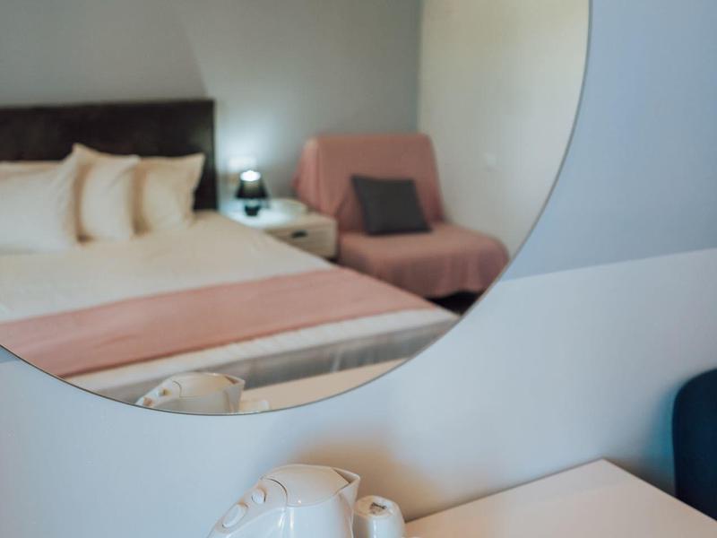 Riflesso in uno specchio rotondo di una camera d'albergo con letto, lampada da comodino e una poltrona rosa.