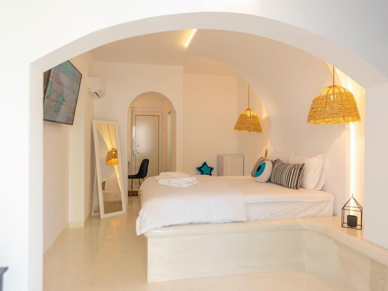 Camera d'hotel luminosa con letto matrimoniale, cuscini, specchio a parete e lampada sospesa in vimini sotto soffitto ad arco.