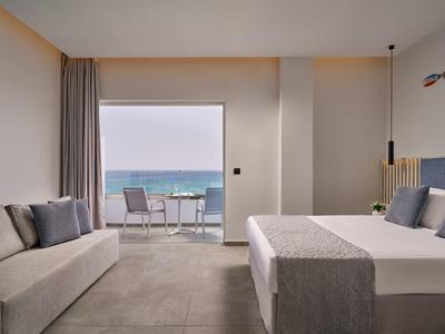 Modern hotel room with sea view, cozy bed, sofa, and balcony with table and chairs.
