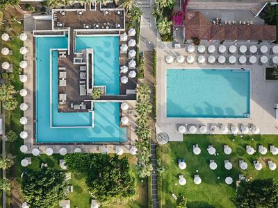 Drone view of two hotel pools with surrounding sun loungers and green areas.