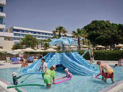 Children play with water fountains in a shallow pool in front of a hotel building under clear skies.