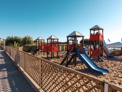 Playground with red and blue equipment on the beach, wooden fence, and sea view.