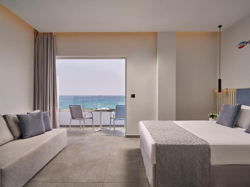 Modern hotel room with sea view, cozy bed, sofa, and balcony with table and chairs.