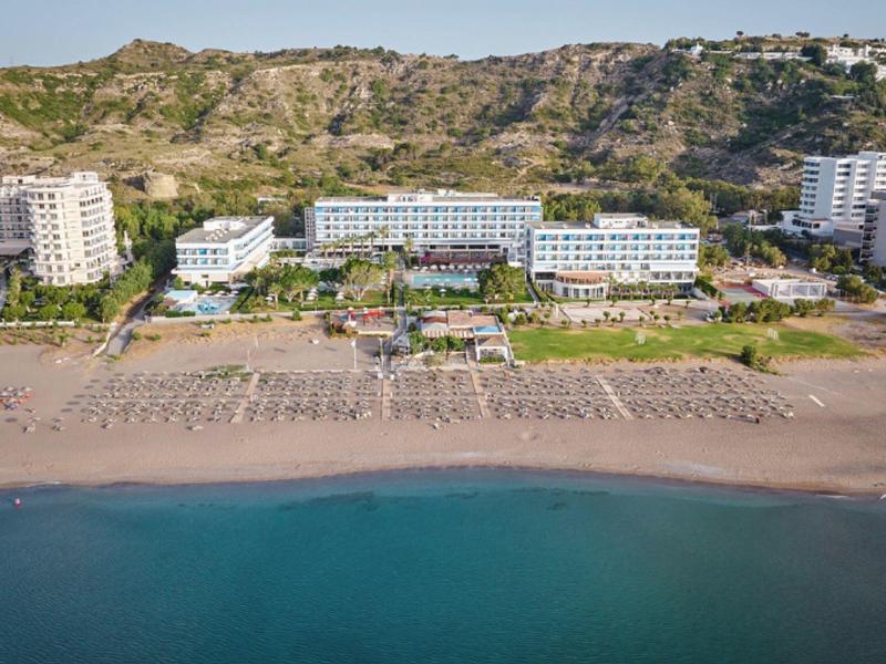 Hotel complex on the beach with sun umbrellas and calm sea in front of a rocky hillside.