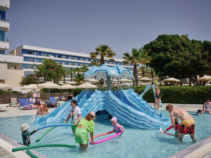 Children play with water fountains in a shallow pool in front of a hotel building under clear skies.