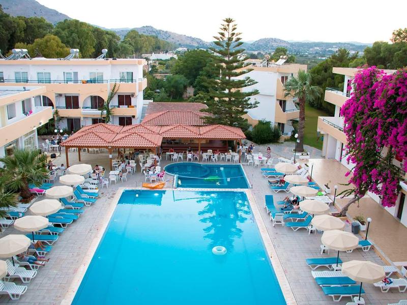 Hotel s velkým bazénem, lehátky a slunečníky, obklopený vícepodlažními budovami a světlými rostlinami.