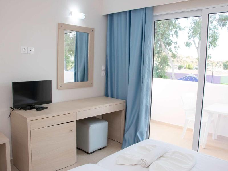 Světelný hotelový pokoj s psacím stolem, televizí, zrcadlem a přístupem na balkon s modrými závěsy.