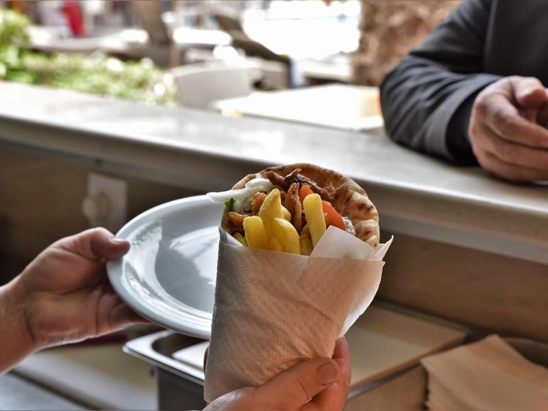 Ruka drží kebab s hranolkami u fast food přepážky, osoba čeká v pozadí.