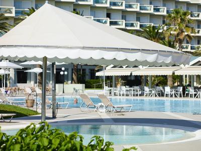 Area piscina moderna dell'hotel con lettini e ombrelloni davanti a un edificio su più piani.