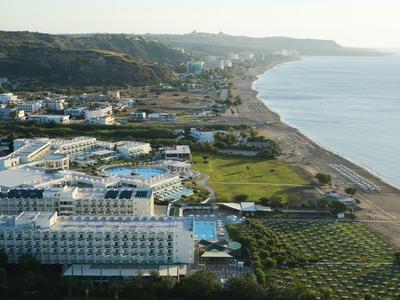 Hotel costiero con edifici bianchi accanto a una spiaggia e uliveti sotto colline boschive.