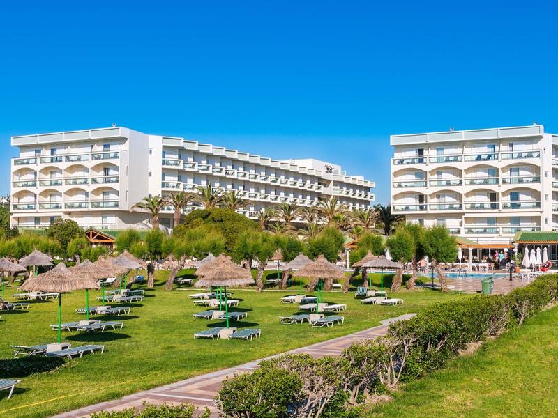 Velká hotelová budova s ​​slunečníky a lehátky na zeleném trávníku pod jasně modrou oblohou.