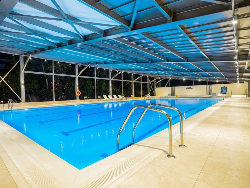 Vytápěný bazén pod modrou skleněnou střechou s kovovým žebříkem a okolními béžovými dlaždicemi.