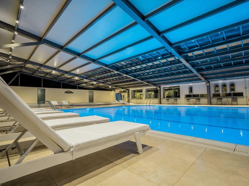 Vnitřní bazén s modrým osvětlením a bílými lehátky pod skleněnou střechou v hotelu.