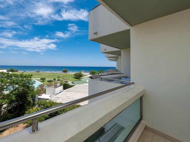 Balkon s proskleným zábradlím a výhledem na moře za jasné oblohy a zelených stromů.